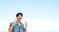 Young Asian male wearing denim shirt making peace signs and smiling against light blue background, expressing positivity and joy for lifestyle and portrait use.