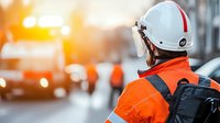 Construction worker in bright orange safety vest and white helmet observing urban worksite during sunset, back view with blurred city background and warm lighting.