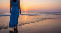 Woman in blue dress walking on beach at sunset with golden light reflecting on wet sand and ocean waves creating peaceful vacation moment.
