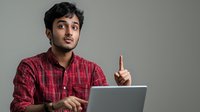Young South Asian professional in red plaid shirt raises index finger while working on laptop, expressing idea or solution against neutral background. Great for business or education concepts.