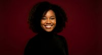 Young African American woman with curly hair smiling warmly in black turtleneck against burgundy background for professional headshot and portrait use.