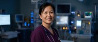 Asian female healthcare professional wearing maroon scrubs and stethoscope smiling in modern hospital monitoring room with illuminated medical equipment and screens in background.
