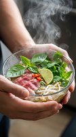Hands holding steaming bowl of Asian noodle soup with fresh basil, lime, red meat and herbs for restaurant, culinary, food styling and cooking content.
