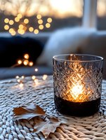 Glowing candle in textured glass holder on woven placemat with autumn leaf and soft bokeh lights creating cozy evening atmosphere. Romantic home decor setting.