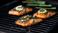 Grilled salmon fillets with lemon slices and fresh herbs cooking on barbecue grates. Juicy fish steaks with caramelized crust and asparagus spears for dinner.