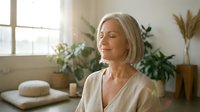 Peaceful mature Caucasian woman with blonde hair meditating in bright wellness spa room with plants and natural light for relaxation and mindfulness.
