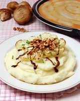Creamy mashed potatoes topped with roasted cauliflower florets, chopped walnuts, and balsamic glaze on white plate, with whole mushrooms in background.