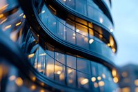 Modern curved glass building facade with warm glowing windows at dusk, creating abstract architectural pattern with bokeh lights and reflections.