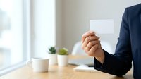 Businessman in dark suit holding blank white card at wooden desk with coffee cups and notebook, bright window background for corporate branding and business communication.