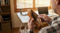 Senior man checking empty wallet at home office desk showing financial stress and money problems during retirement planning.