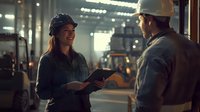 Female Asian industrial engineer in protective helmet discussing project details with construction worker at manufacturing facility during evening shift.