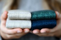 Hands holding colorful metallic thread spools in silver, teal and navy blue colors against blurred background, showing textile crafting supplies.