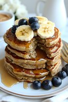 Stack of fluffy banana pancakes topped with fresh blueberries, sliced bananas and maple syrup drizzle on white plate, with bite taken showing texture.