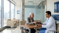 Professional doctor consulting with mature female patient in modern medical office with city view windows and contemporary furniture.