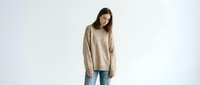Young woman with long dark hair and medium skin tone wearing a casual beige oversweater and blue jeans, standing against white background for fashion and lifestyle content.