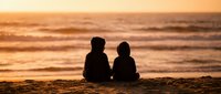 Two children sitting together on beach sand watching golden sunset over ocean waves creating peaceful silhouette moment for family vacation memories.