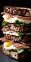Gourmet breakfast sandwich with fried eggs, ham, and fresh greens layered between slices of artisanal multigrain bread against dark background.