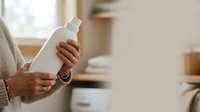 Senior woman holding white cosmetic bottle with pump dispenser in home interior setting, skincare product presentation.