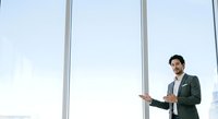 Young male professional with dark hair and beard, wearing olive green blazer and white shirt, gesturing while speaking in modern office with floor-to-ceiling windows, suitable for business presentations and corporate communications.