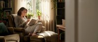 Middle-aged man with light skin tone reading tablet while relaxing in comfortable armchair by sunny window in cozy home library setting with bookshelves.