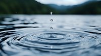 Water drop falling into calm lake surface creating concentric ripple circles against blurred mountain landscape background in moody blue tones.