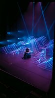Person sitting amid glowing neon wireframe waves and blue laser beams in dark futuristic environment. Digital art installation with interactive light display.