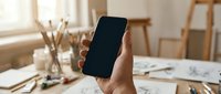 Hand holding smartphone with blank black screen in bright creative workspace with art supplies and sketches on desk for mockup presentations.