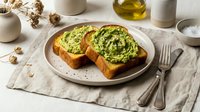 Avocado toast with herbs on sourdough bread served on ceramic plate with olive oil and natural linen table setting for healthy breakfast or brunch meal.