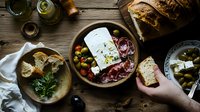 Mediterranean appetizer platter with feta cheese, olives, bread, and cured meats served on rustic wooden table with olive oil bottles and fresh herbs for sharing.