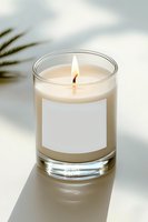 Burning aromatic candle in glass container with blank label on white surface, soft palm leaf shadow creates minimalist spa atmosphere. Suitable for branding mockup.