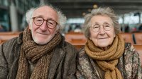 Happy elderly couple wearing warm scarves smiling together in cozy indoor setting. Senior man and woman with glasses enjoying retirement lifestyle.