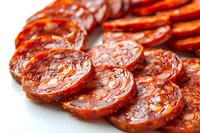 Sliced spicy chorizo sausage with red paprika coating arranged in rows on white background, showing texture and marbling of cured meat. Close-up macro food photography.