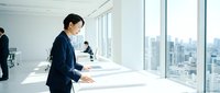 Asian female professional in dark business suit working at white desk in modern high-rise office with city skyline view, corporate workspace environment.
