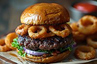 Juicy beef burger topped with crispy onion rings on brioche bun, served with lettuce, red onion and side of golden fried rings. Fresh restaurant meal.