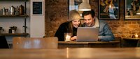 Young couple enjoying coffee while using laptop together in cozy cafe with exposed brick walls and warm lighting atmosphere for remote work or leisure.