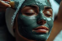 Young woman receiving luxury spa facial treatment with green clay mask being applied with brush, eyes closed in relaxation, close-up beauty shot with soft lighting.
