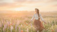 Woman with curly blonde hair wearing white blouse and brown skirt standing in blooming lupine field at golden hour sunset, ideal for lifestyle and nature editorial use.