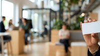 Hand holding blank white card in modern office lobby with blurred colleagues and green plants in background, suitable for business messaging and corporate communications.