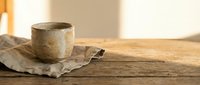 Rustic ceramic cup on linen napkin atop weathered wooden table with warm sunlight, suitable for home decor and lifestyle content.