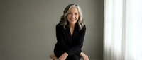Confident mature Caucasian businesswoman with gray hair smiling in black blazer sitting by window in modern office for corporate professional headshot.