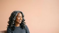 Middle-aged Black woman with long wavy gray and dark hair, wearing a charcoal ribbed sweater, smiling while looking to the side against a warm beige background.