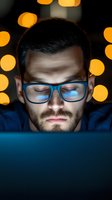Young Caucasian male with glasses illuminated by screen light at night, bokeh lights in background, focused expression while working late on computer device.