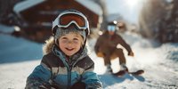 Happy young child in ski goggles and winter jacket smiling on snowy mountain slope with skier in background during sunny winter day at resort.