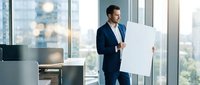 Professional businessman in navy suit holding blank white poster in modern office with city view through large windows for corporate presentation mockup.