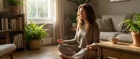 Woman with long curly brown hair, Caucasian ethnicity, sitting in lotus position on wooden floor in bright living room, meditating with eyes closed, wearing beige cardigan. Peaceful home wellness scene.