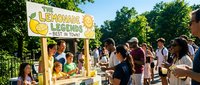 Outdoor lemonade stand with yellow striped awning serving refreshing drinks to diverse customers on sunny day in park setting.