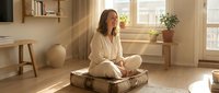 Young woman meditating in peaceful home interior with natural sunlight streaming through windows. Mindful wellness lifestyle concept for relaxation.