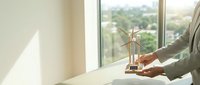 Businessman hands holding miniature wind turbine model on office desk near window with city view representing renewable energy investment and sustainability.