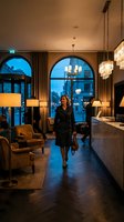 Elegant businesswoman in dark coat walking through luxury hotel lobby with arched windows and warm evening lighting for hospitality and travel concepts.