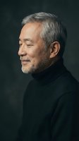 Mature Asian man with gray beard smiling in profile wearing black turtleneck sweater against dark background for portrait photography and lifestyle content.